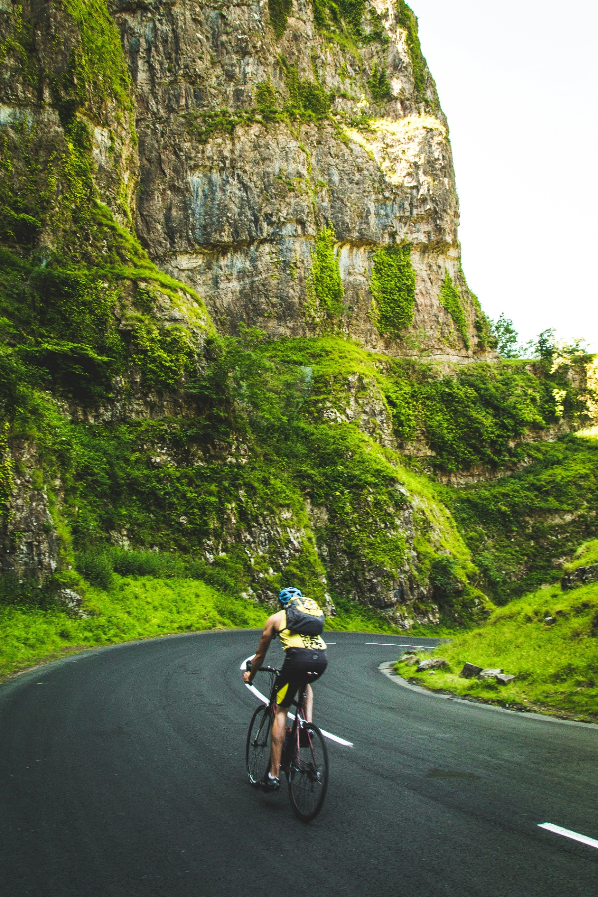 Road cyclist riding along a winding road beneath a towering green cliff